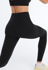 Osoba v čiernych legínach s vysokým pásom a bielych teniskách v póze výpadu na jednoliatom bielom pozadí.