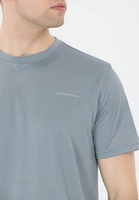 Vaaleanharmaa urheilupaita, joka on valmistettu sileästä, kosteutta siirtävästä kankaasta. Siinä on lyhyet hihat ja huomaamaton "ENDURANCE"-logo rinnassa.