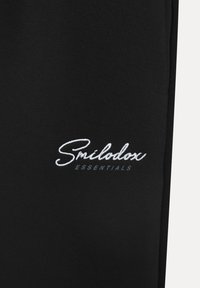 Černé tepláky jsou vyrobené z měkké bavlny a mají vyšívané logo "Smilodon Essentials" v bílé a šedé barvě.