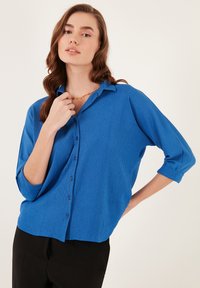 Camisa azul de botón con un acabado texturizado, mangas enrolladas y un corte relajado. Combinada con pantalones negros.