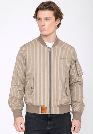 Junger Mann trägt eine beige Bomberjacke mit feinen Nadelstreifen, Reißverschluss vorne und Taschen, steht vor einem schlichten weißen Hintergrund.