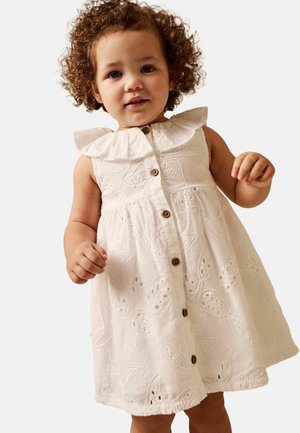 Tout-petit aux cheveux bouclés portant une robe blanche brodée sans manches avec des boutons et un col à volants, debout devant un fond uni.