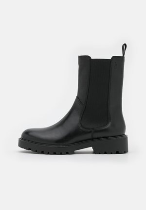 Bottine en cuir noir avec panneaux latéraux en élasthanne, bout rond, semelle épaisse basse et languette en haut pour faciliter l'enfilage.