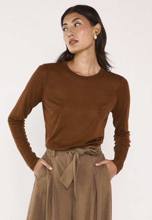 Donna con capelli scuri che indossa un top marrone a maniche lunghe e pantaloni beige a vita alta con cintura annodata, mani nelle tasche, guarda di lato.