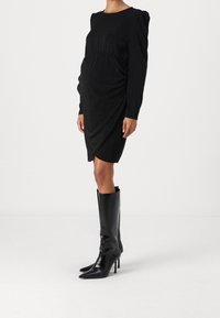 Robe noire à manches longues avec une taille cintrée, un design drapé et un léger scintillement. Associée à des bottines noires à talons hauts.