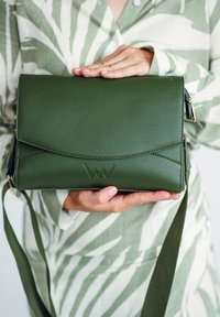 Zelená kožená kabelka s klopou, embosovaným logem a nastavitelným popruhem. Hladký povrch s ostrými designovými prvky.