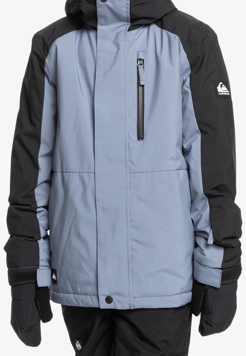 Veste d'hiver isolante bleu clair et noire avec poche poitrine zippée, capuche, et gants noirs portés par une personne sur fond blanc.