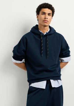 Junger Mann mit lockigem Haar, der einen dunkelblauen Hoodie über einem gestreiften Hemd trägt und mit den Händen hinter dem Rücken an einer schlichten hellen Wand steht.