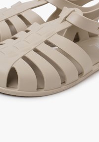 Sandalias de plástico beige con un diseño de correas, que cuentan con múltiples ranuras abiertas para la ventilación y una suela plana. Construcción duradera y ligera.