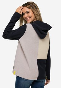 La felpa con cappuccio presenta un mix di pannelli beige, blu navy e grigio chiaro, un tessuto testurizzato e un accento giallo a contrasto sull'orlo.