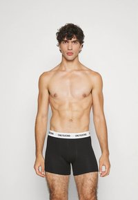 Zwarte boxershorts met een witte tailleband waarop "ONLY & SONS" staat. Het materiaal lijkt glad en sluit nauw aan rond de taille en dijen.