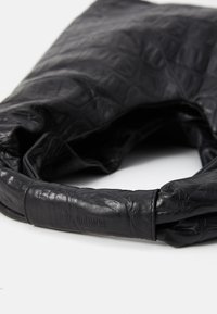 Bolso de mano de cuero negro con un acabado texturizado, un diseño suave y plegable, y un asa lisa y redondeada. Presenta un logo grabado en la correa.