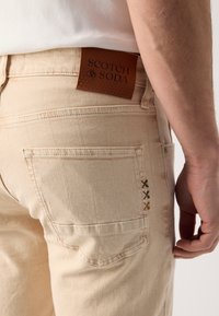 Jeans in cotone beige con etichetta in pelle SCOTCH & SODA, dotati di una tasca con accenti di ricamo colorato e design a gamba dritta.