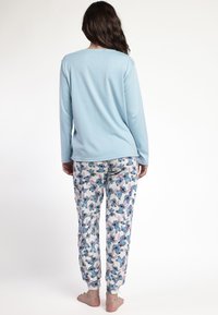 Mujer de pie descalza, de espaldas, vistiendo una blusa de manga larga azul claro y pantalones de pijama blancos con estampado de perrito azul.