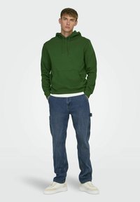Felpa verde con tasca frontale e cappuccio regolabile, abbinata a pantaloni cargo in denim blu, dotati di tasche laterali. Scarpe da ginnastica bianche completano il look.
