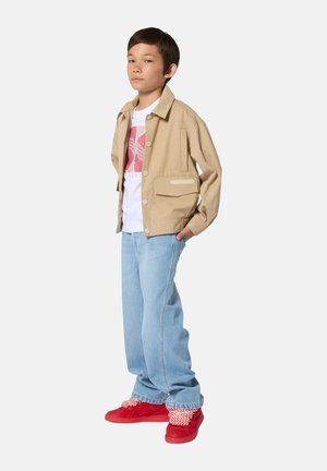 Jongen staat met handen in zijn zakken, draagt een beige jas, wit shirt met print, lichtblauwe spijkerbroek en rode sneakers met gemêleerde sokken.