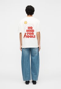 Λευκό βαμβακερό t-shirt με κόκκινο γραφικό κείμενο «NO NEED FOR TOOLS» και συνοδευτικές απλές εικονογραφήσεις στην πλάτη, συνδυασμένο με φαρδιά μπλε τζιν.