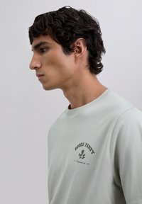 Junger Mann mit dunkelbraunen lockigen Haaren trägt ein hellgraues T-Shirt mit dem Aufdruck "Rodeo Party" und einem floralem Grafikdesign auf der Brust. Er schaut nach links.