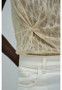 Close-up van een beige gebreide top met gedraaide voorkant, gecombineerd met hoge witte denim broeken op een model met een donkere huid.