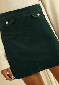 Zelená kordurová sukně s dvěma předními kapsami a měděnými knoflíky, rovný střih a měkká textura. Lem sukně sahá nad kolena.