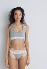 Szara bawełniana sportowa biustonosz z białą opaską z logo Calvin Klein oraz dopasowanym szarym thongiem. Model ma jasną karnację z widocznymi wariacjami w strukturze skóry.