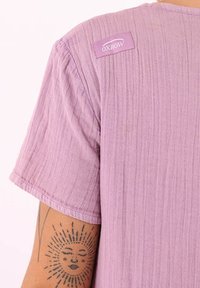 Top lilas à manches courtes en tissu texturé, avec une étiquette qui indique "OXBOW" sur l'épaule. Couleur uniforme et simple, sans motifs.