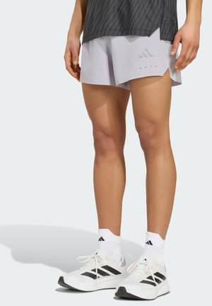 Person trägt hellgraue Adidas-Shorts, weiße Adidas-Socken und weiße Laufschuhe mit schwarzen Streifen und steht auf hellem Hintergrund.