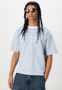 T-shirt à manches courtes rayé en bleu clair et blanc avec des accents bleu marine, fait de tissu doux. Associé à un short en denim, porté avec des lunettes de soleil.