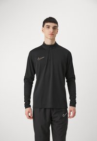 Zwarte Nike langarm top met een ritssluiting bij de kraag. Bevat een gouden Nike-logo op de linkerborst, een gladde stof en een aansluitend ontwerp.