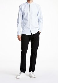 Camicia a righe azzurro chiaro con colletto alla francese, abbinata a pantaloni neri e sneakers bianche, in piedi contro uno sfondo neutro.