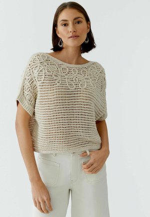 Frau trägt ein beiges, gehäkeltes Kurzarm-Top und helle Hosen, posiert mit einer Hand in der Tasche vor neutralem Hintergrund.