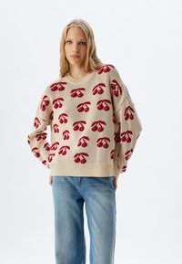 Beige gebreide trui met een rood kersenpatroon, brede mouwen en een geribde zoom. Gepaard met blauwe jeans.