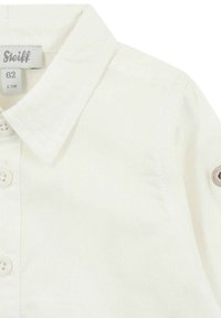 Camicetta bianca in cotone con colletto, dotata di bottoni frontali e un'etichetta sul colletto. Un dettaglio significativo è il patch con logo sul maniche.