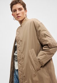 Μπεζ μπουφάν bomber με ραφές σε σχήμα καπιτονέ, κλείσιμο με κουμπιά πάτου, πλαϊνές τσέπες και τσέπη με φερμουάρ στο μανίκι. Λείο ύφασμα και άνετη γραμμή.