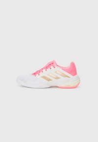 Zapatilla deportiva blanca con acentos en rosa, que presenta rayas doradas, una parte superior texturizada y una suela acolchada. Cordones en color rosa a juego.