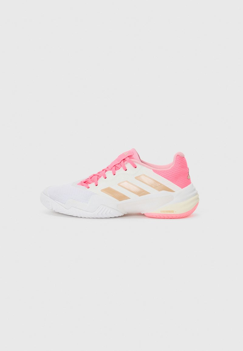 Zapatilla deportiva blanca con acentos en rosa, que presenta rayas doradas, una parte superior texturizada y una suela acolchada. Cordones en color rosa a juego.