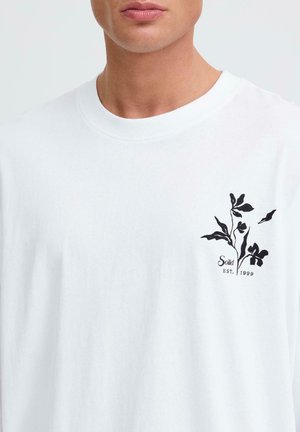 Man wearing white crewneck shirt with black floral graphic and text "Solid EST. 1999" on left chest.