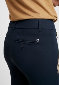 Marinblå, figursydda byxor i stretchtyg som har en bakficka med en svart knapp och en metalllogotyp nära fickans söm.