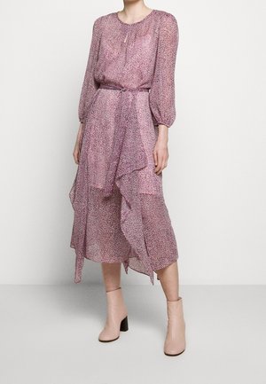 Kobieta ubrana w różową, wzorzystą, długorękawową sukienkę midi z falbanami i wiązaniem w talii, w połączeniu z beżowymi botkami za kostkę.