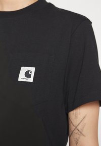 T-shirt noir en coton avec une poche poitrine portant un logo Carhartt blanc. Manches courtes et col rond standard avec une texture douce.