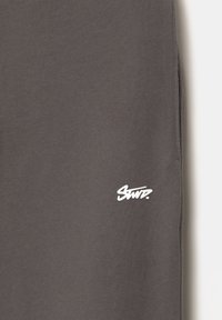 Tissu gris foncé avec un petit logo blanc stylisé près du bord, montrant la texture et les détails de la couture sur le côté droit.