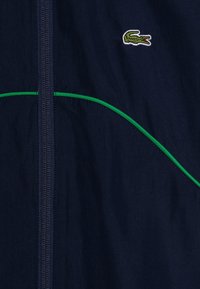 Chaqueta azul marino con cremallera frontal, detalle de ribete curvado verde y un pequeño logo bordado de cocodrilo verde en el pecho.