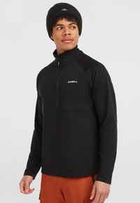Zwarte fleece pullover met een kwartrits en hoge kraag, voorzien van een wit logo, gecombineerd met oranje broek en een zwarte muts. Glad textuur en aansluitend ontwerp.