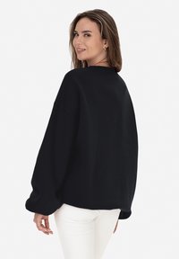 Sudadera negra de gran tamaño con mangas largas y caídas, y un cuello de canalé. Tejido suave, costuras mínimas y un corte relajado.