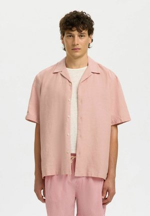 Mlad moški s kratkimi kodrastimi lasmi, oblečen v svetlo rožnato srajco s kratkimi rokavi čez belo majico in hlače z rožnatimi črtami.