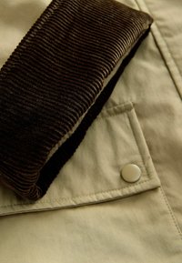 Tessuto beige con texture visibile, caratterizzato da un corduroy marrone scuro ribattuto. Include un dettaglio a bottone sulla tasca.