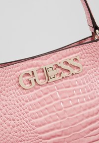 Borsa rosa con texture a coccodrillo, caratterizzata da un prominente logo dorato "GUESS" e una finitura in vernice liscia sulla superficie.