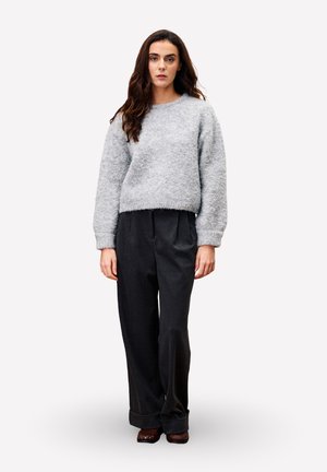 Femme aux longs cheveux noirs portant un pull en laine léger gris, un pantalon large noir et des chaussures marron, se tenant devant un fond neutre.