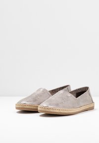 Un par de bailarinas de esparto plateadas y grises con adornos de yute trenzado y suelas de goma sobre una superficie blanca.