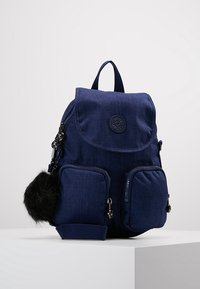Sac à dos en tissu bleu marine avec une fermeture à rabat, deux poches zippées à l'avant et un logo rond. Il est orné d'un pompon en fourrure sur le côté.